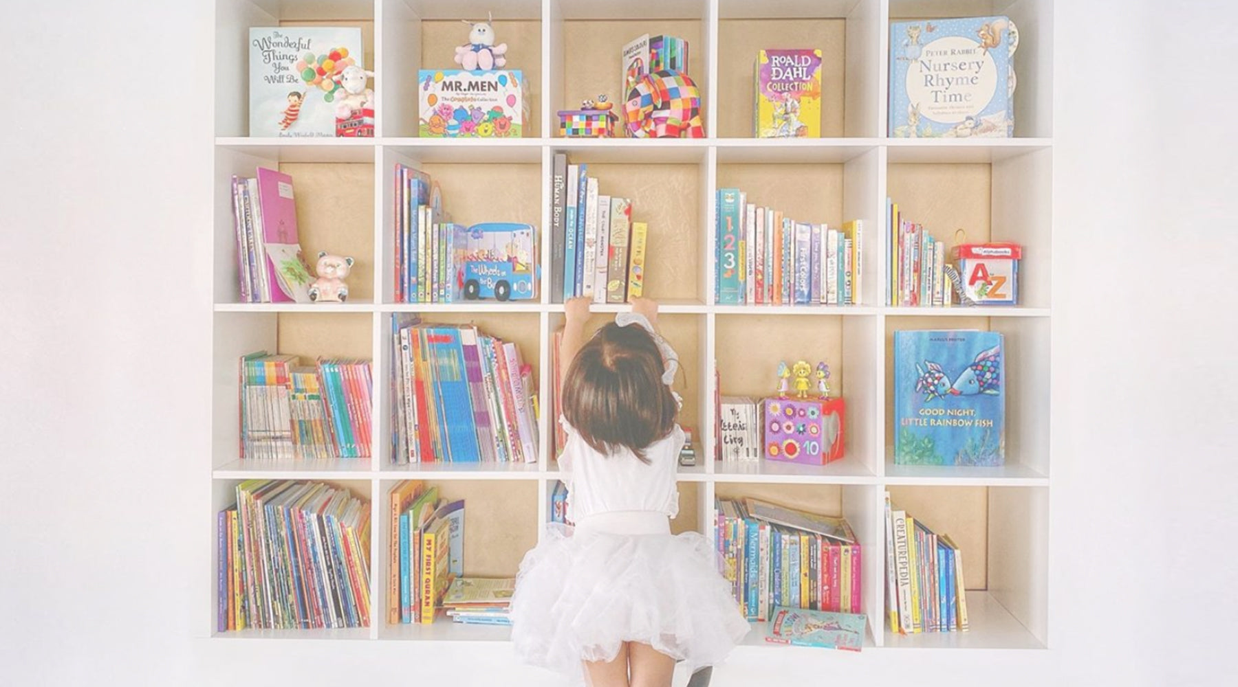 Little girl in white dress reaching for books in her bookshelf